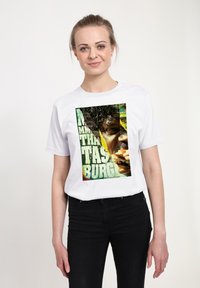 Bílá bavlněná tričko s barevným grafickým potiskem tváře osoby, která jí hamburger, s odvážným textem a živými pozadími.