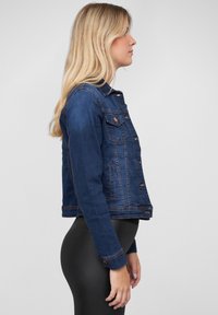 Veste en denim bleu foncé, avec boutons à l'avant, deux poches poitrine et accents cousus. Coupée à la longueur des hanches avec des manches longues.