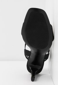Sandale noire à talon haut avec semelle texturée et talon fin, inclinée contre un mur d’angle blanc et une surface de sol.
