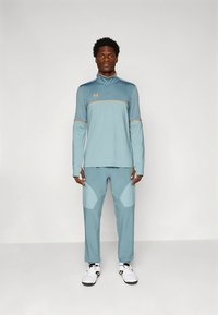 Világoskék sportos ruha hosszú ujjakkal, cipzáras nyakkal és színes blokkokkal díszített nadrággal, világosabb kék díszítéssel. Fekete-fehér cipők.