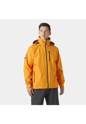 Man met een feloranje gewatteerde jas met capuchon, zwarte ritszakken en logo's op de borst en mouw, staand tegen een lichte achtergrond.