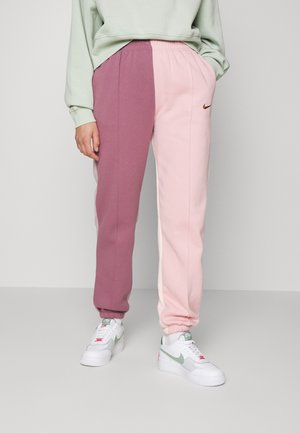 Personne portant un jogging bicolore avec une jambe rose poussière et l'autre rose clair, associé à des baskets blanches et un sweat léger.