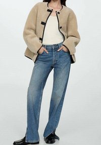 Giacca in pile beige con dettagli neri e chiusure a cordoncino, abbinata a jeans a gamba larga blu chiaro e scarpe nere, mani nelle tasche.