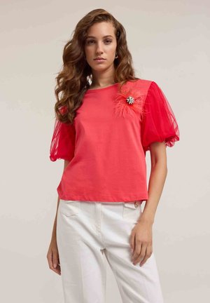 Rode katoenen t-shirt met korte, doorschijnende tule pofmouwen. Heeft een decoratieve veren- en strass-steentjesversiering op de linker schouder.