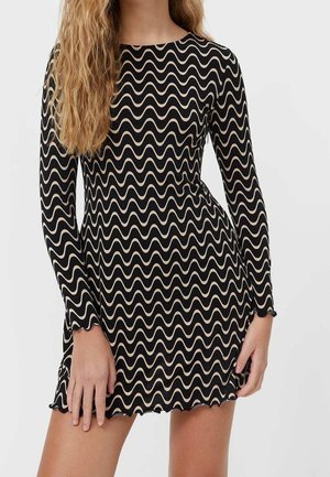 Vrouw draagt een zwart mini-jurkje met lange mouwen en een beige golvend lijnpatroon, staand met handen nabij de heupen tegen een eenvoudige achtergrond.