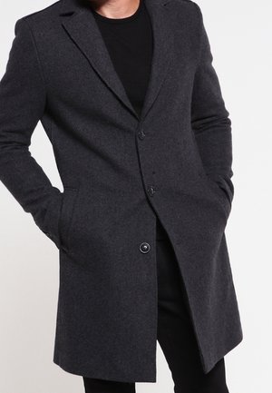 Homme portant un manteau en laine gris foncé boutonné, les mains dans les poches, associé à un pantalon noir et une chemise noire.