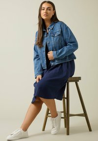 Ljusblå denimjacka, kort design med fickor; kombinerad med en navyblå klänning och vita sneakers. Modell sitter på en träpall.