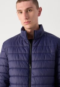 Marineblå quilted puffer-jakke med høy krage, glidelås foran og horisontale sømmer; glatt tekstur og lett utseende.