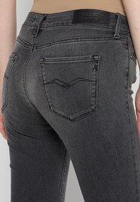 Gros plan d'une personne portant un jean Replay noir délavé avec des poches arrière et une étiquette en cuir de la marque sur la ceinture.