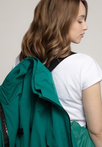 Türkisfarbene Jacke mit einer glatten, matten Oberfläche, gehalten über einem weißen T-Shirt. Die Jacke hat einen Reißverschluss und einen schwarzen Riemen. Welliges Haar ist sichtbar.