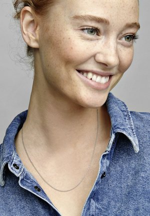 Collana d'argento al collo, con una catena delicata. Il modello indossa una camicia di jeans blu con colletto a punta e bottoni scuri.
