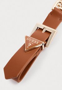 Bracelet en cuir marron avec une boucle en laiton doré en forme de triangle portant un logo. Texture lisse avec une extrémité effilée, les détails incluent un fermoir.