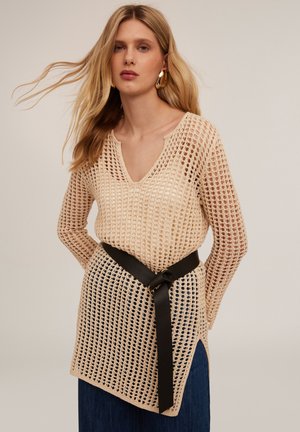 Beige gehaakte trui met een V-hals en lange mouwen, voorzien van een zwarte stoffen riem die om de taille is gestrikt en zijsplitten voor extra detail.