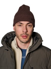Mann mit leichtem Bart und Nasenring, der eine braune Strickmütze und eine geschichtete Jacke mit beigem Fleece und blauem Hemd darunter trägt, blickt nach vorne.