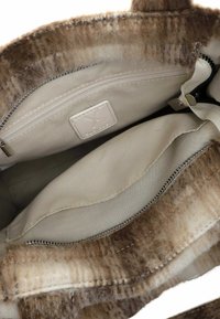 Interior de una bolsa de tela beige que muestra un bolsillo con cremallera, una etiqueta y un borde exterior marrón suave y aterciopelado.