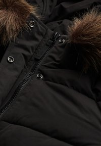 Sort pufferjakke med lynlåslukning med sølvhardware og aftagelig faux fur-pels på hætten, med en quiltet tekstur.