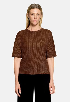Vrouw met steil blond haar, gekleed in een bruin gebreid trui met korte mouwen en een zwarte broek, staande tegen een effen lichte achtergrond.