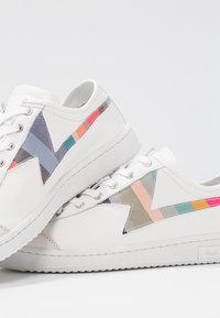 Baskets basses blanches avec des rayures en zigzag multicolores sur les côtés et des lacets blancs, présentées superposées sur un fond clair.