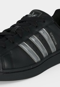 Μαύρο sneaker Adidas Superstar με ασημί ρίγες, μαύρα κορδόνια και λογότυπο στο πλάι και τη γλώσσα.