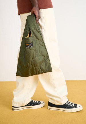 Quilted green tote bag with a handle, featuring a label. Paired with cream-colored pants and navy sneakers on a yellow floor.