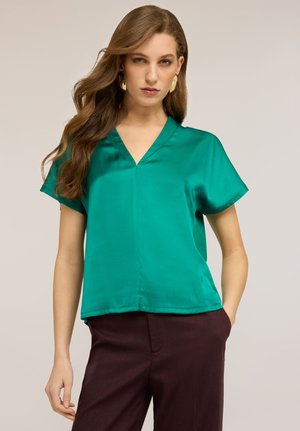 Kobieta z długimi brązowymi włosami, nosząca złote kolczyki, błyszczącą zieloną bluzkę z krótkim rękawem i dekoltem w kształcie litery V oraz ciemnobrązowe spodnie, z jedną ręką w kieszeni.