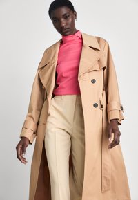 Beige trenchcoat met dubbele knopen, brede revers en lange mouwen, gedragen over een roze coltrui en crème broek.