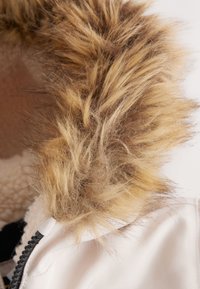Faux fur hood trim in shades of brown and blonde, attached to a white outerwear garment. Features soft, thick texture and detailed stitching.