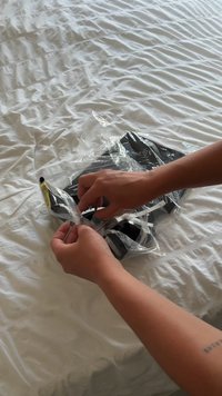 Rokas atver caurspīdīgu plastmasas iepakojumu, kurā atrodas melna sejas maska uz baltas teksturētas gultas veļas.