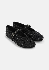 Zapatos negros con punta redondeada, que presentan una tela texturizada adornada con tachuelas elevadas y una correa con hebilla a través del empeine.