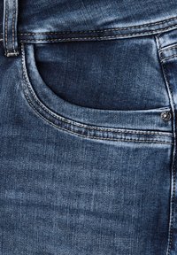 Jeans de mezclilla con un lavado azul oscuro, que cuentan con un bolsillo frontal, costuras visibles y un acento de remache metálico en el bolsillo. Textura suave.