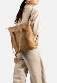 Zaino in pelle color tan con chiusura a ribalta, spallacci regolabili e finitura testurizzata. Presenta un'etichetta con il marchio e poggia su un outfit in tessuto leggero.