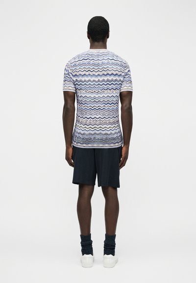 Άνδρας που φοράει κοντομάνικο πουκάμισο με πολύχρωμο σχέδιο ζιγκ ζαγκ, μαύρο σορτς, σκούρες κάλτσες και λευκά αθλητικά παπούτσια, στέκεται γυρισμένος με την πλάτη.