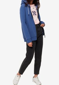 Woman wearing blue zip-up hoodie, pink graphic t-shirt, black high-waist jeans, and white sneakers, standing with one hand in pocket.