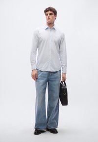 Vaaleansininen slim fit -kauluspaita yhdistettynä leveälahkeisiin sinisiin farkkuihin. Malli pitää mustaa nahkalaukkua ja on pukeutunut mustiin kenkiin.