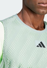 Una camiseta sin mangas deportiva de color verde claro con textura de malla, que presenta un escote redondeado y patrones de puntos. Incluye un parche del logo de Adidas en negro.