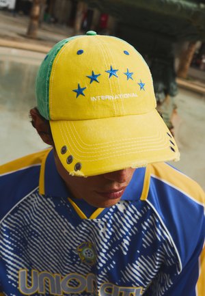 Personne portant une casquette jaune et verte avec des étoiles bleues et le texte "INTERNATIONAL", ainsi qu'un maillot de sport bleu et jaune avec un design à motifs.