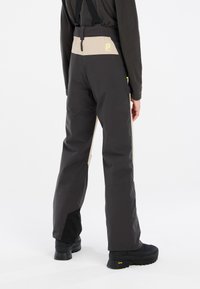 Pantalons imperméables noirs et beiges avec bretelles ajustables, une texture lisse et des zip sur les côtés pour la ventilation et les poches.