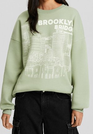 Nainen, joka pukeutunut vaaleanvihreään collegepaitaan, jossa valkoinen Brooklynin siluetti ja teksti "WELCOME", yhdistetty mustiin cargo-housuihin.