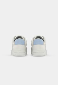Hvite lavtopp sneakers med lyseblå hælpatcher, merkevarenavnet "Tommy Hilfiger" preget på baksole, på en ensfarget bakgrunn.