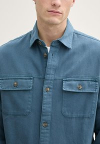 Cămașă din denim albastru cu guler ascuțit, două buzunare pe piept cu închideri cu nasturi și un material texturat. Nasturii sunt maro.