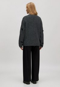 Übergroßer schwarzer und grauer Strickpullover mit langen Ärmeln, der aus strukturiertem Stoff gefertigt ist und ausgeschnittene Akzente an den Ellbogen aufweist, getragen über einer schwarzen Hose.