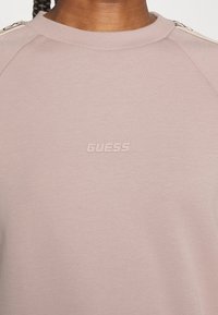 Sweatshirt rose clair avec un col rond et des manches raglan. Présente le logo "GUESS" en relief légèrement texturé, avec des accents métalliques sur les épaules.