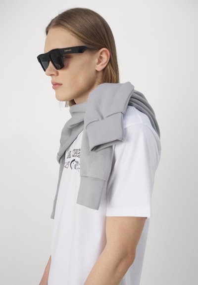 Gray cotton sweater draped over a white t-shirt with graphic print; model wears black sunglasses with gold accents.