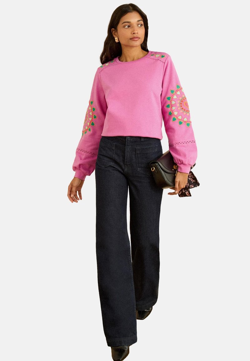 Roze sweatshirt met decoratieve borduurwerk in groen en oranje, gecombineerd met donkere jeans met rechte pijpen. Accessoires omvatten een kleine zwarte handtas.