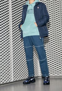 Marinblå isolerad jacka, mintgrön huvtröja med texten "LIMITED", mörkblå byxor med detaljerade sömmar och svart-blå sportskor. Texturerad bakgrund.