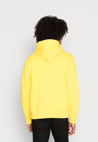 Sudadera amarilla con capucha de cuello alto y cordón ajustable. Tejido liso, puños y dobladillo acanalados. Vista trasera con pantalones negros visibles.