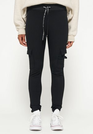 Leggings neri in tessuto elasticizzato con tasche cargo, vita con coulisse e design aderente. Indossati con scarpe da ginnastica bianche.
