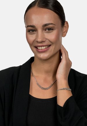 Silver chain necklace and bracelet set, featuring interlocking links with a polished finish. Worn with a black top and jacket.