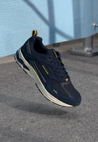 Námořnické modré sportovní boty z prodyšného a syntetického materiálu, s žlutými akcenty a aerodynamickým designem, visící nad šedým povrchem.