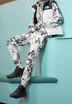 Biała kamizelka puchowa z czarnymi akcentami na tle zestawu w abstrakcyjny czarno-biały wzór. Całość uzupełniają czarne buty sportowe.